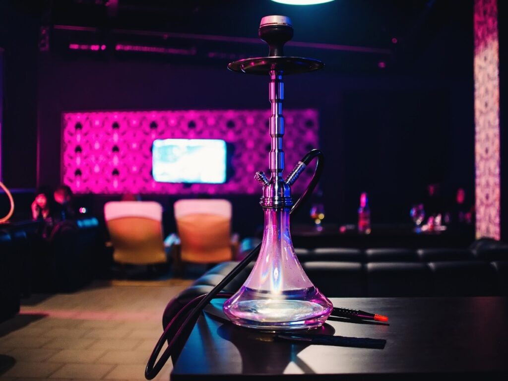Elegant glass hookah pipe with hose on a table in a cozy Hookah Shop Houston lounge, featuring vibrant purple neon lighting, comfortable seating, and a relaxing atmosphere.