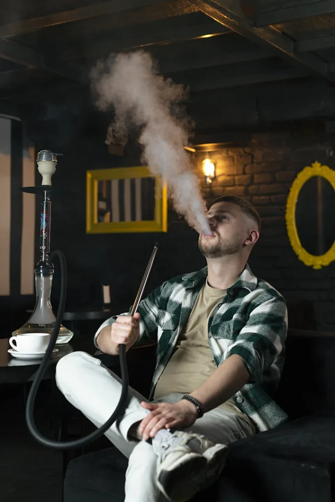 Man smoking hookah with thick smoke in a cozy Hookah Shop Houston lounge with yellow framed mirrors and dim lights.