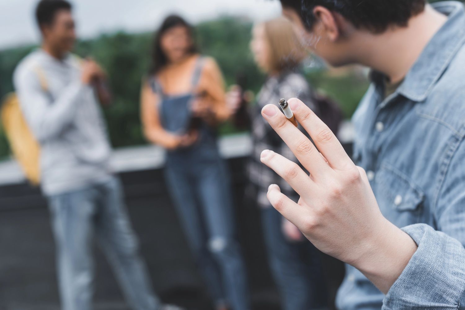 A person holding a lit cigarette outdoors with friends, great for a cigarette shop Broken Arrow.