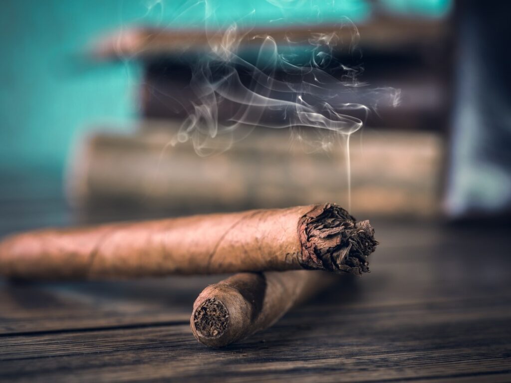 Two intricately crafted cigars resting on a rustic wooden table, with a soft plume of smoke curling upward, offered exclusively by Cigarette Shop Houston.