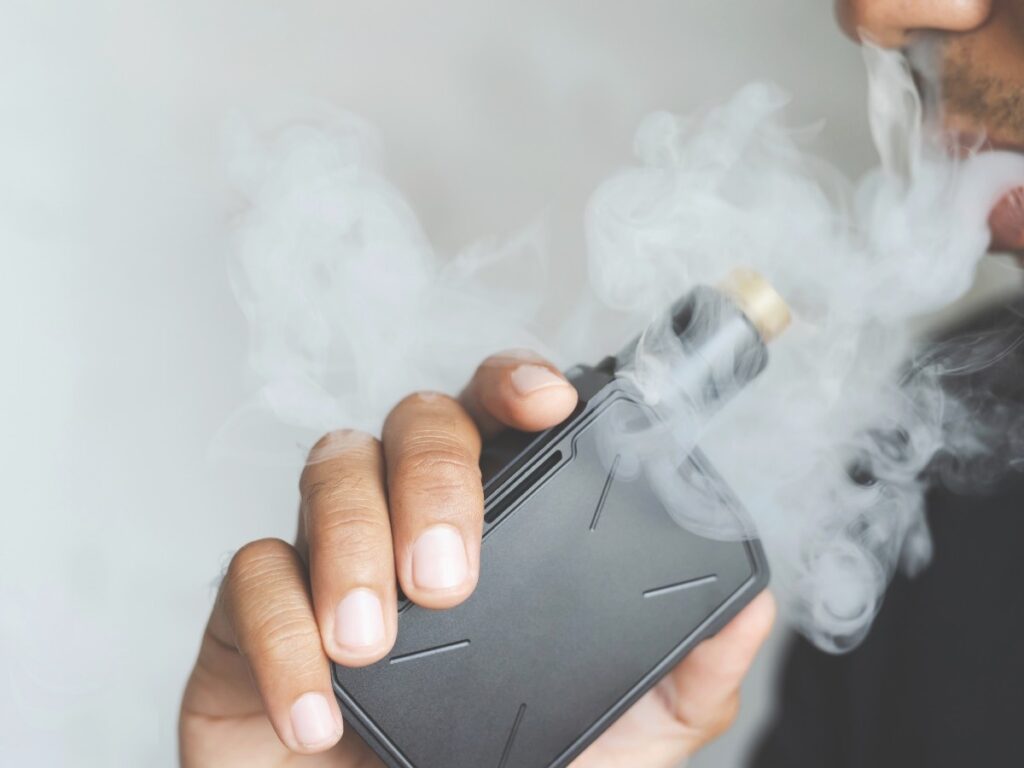 Close-up of a person holding a sleek black vape device with gold tip, exhaling thick vapor cloud in a modern setting at Vape Shop Houston.