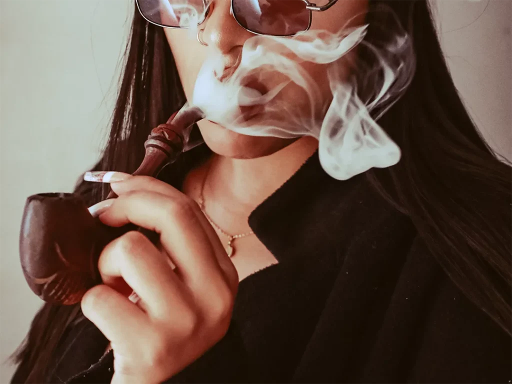 A person smoking a stylish glass pipe in Houston, with smoke swirling, wearing sunglasses and a dark coat, promoting glass pipes Houston.