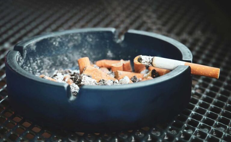 A black ashtray full of cigarette butts with one lit cigarette resting on the rim, sold at your best smoke shop broken arrow.