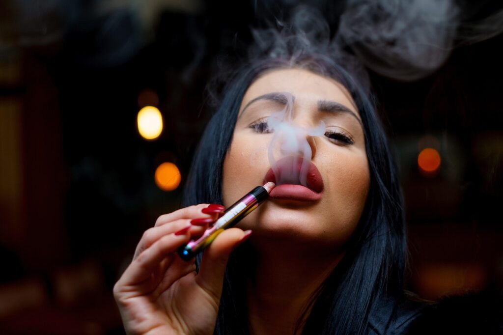 Attractive woman exhaling a cloud of thick vapor from a small, colorful vape pen, get your flavors at e-cigarettes broken arrow.