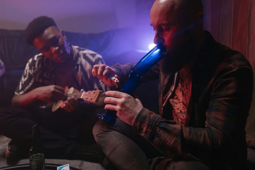 Two men share a blue bong in a dim living room while one plays a ukulele — casual hangout image for cigarette shop Tulsa.