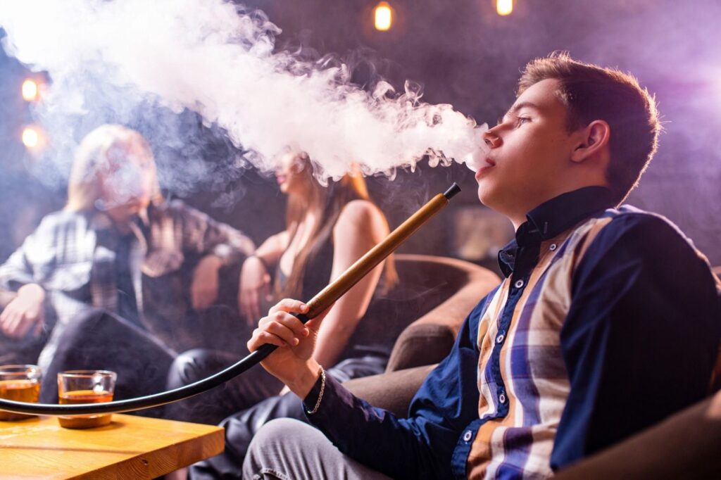 Young man smoking shisha in dim lounge with friends, featured in smoke shop Tulsa campaign for social smoking lifestyle.