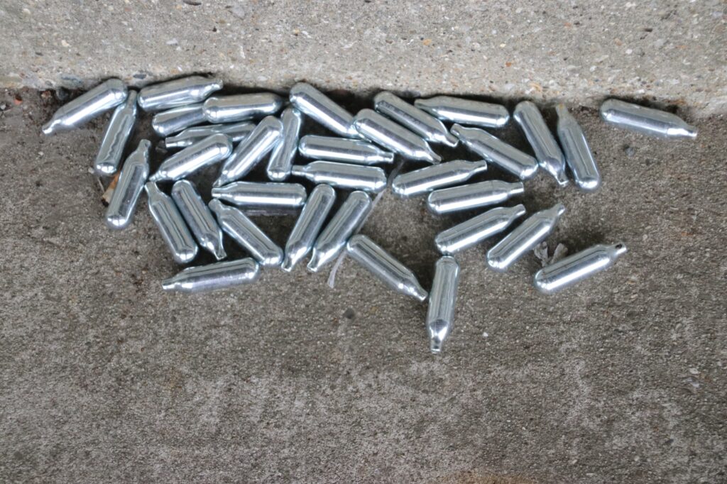 Multiple discarded silver nitrous oxide Owasso canisters lying scattered on a concrete surface near a wall outside.
