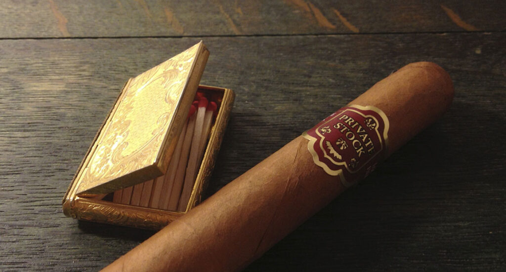 Single private stock cigar and a golden matchbox on a wood surface, essentials for any visit to a great cigar shop owasso.
