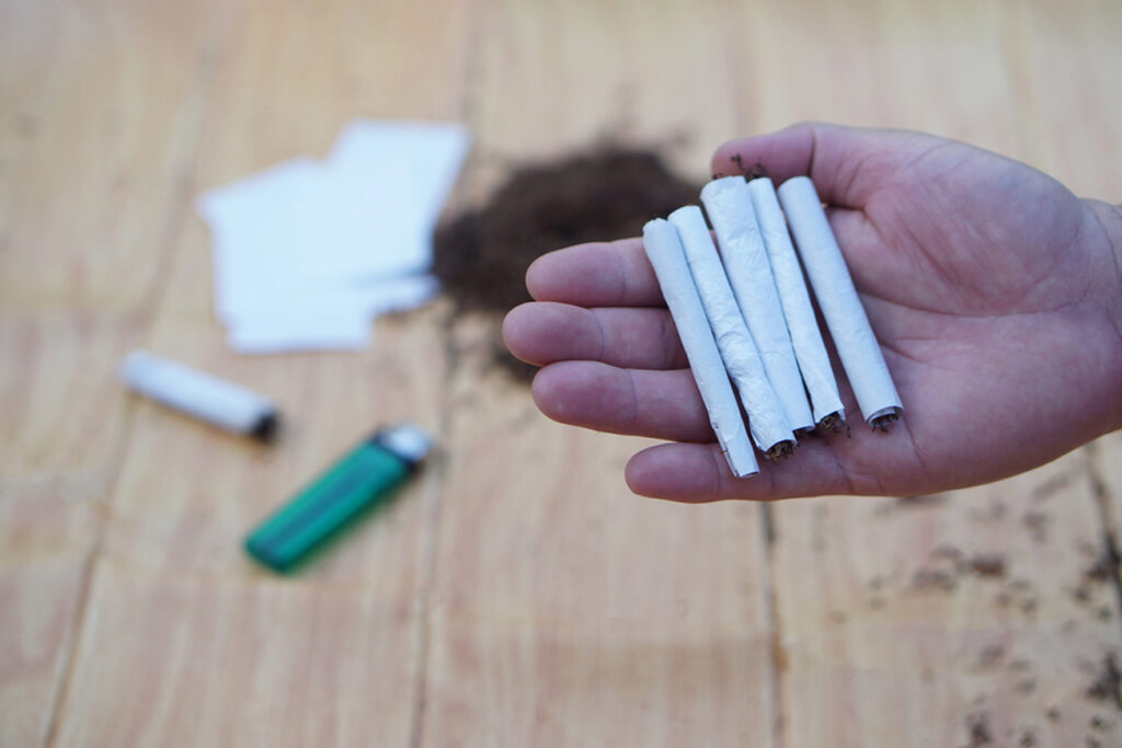 Close-up of a hand holding multiple hand-rolled joints crafted with premium rolling papers Sapulpa, featuring scattered herb, filters, and a green lighter on a wooden table for a natural smoking experience.