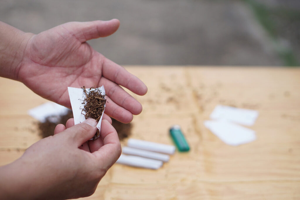 Hands expertly rolling dried herb into thin rolling papers Sapulpa, with completed joints, extra papers, and a lighter nearby on a rustic wood surface, showcasing easy DIY joint preparation.