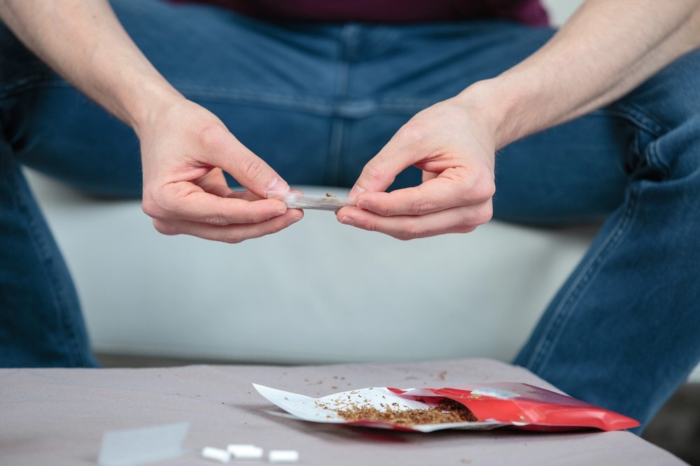 Hands filling a clear cone with loose tobacco from open red pouch on table, premium rolling accessories from local head shop in Sapulpa.