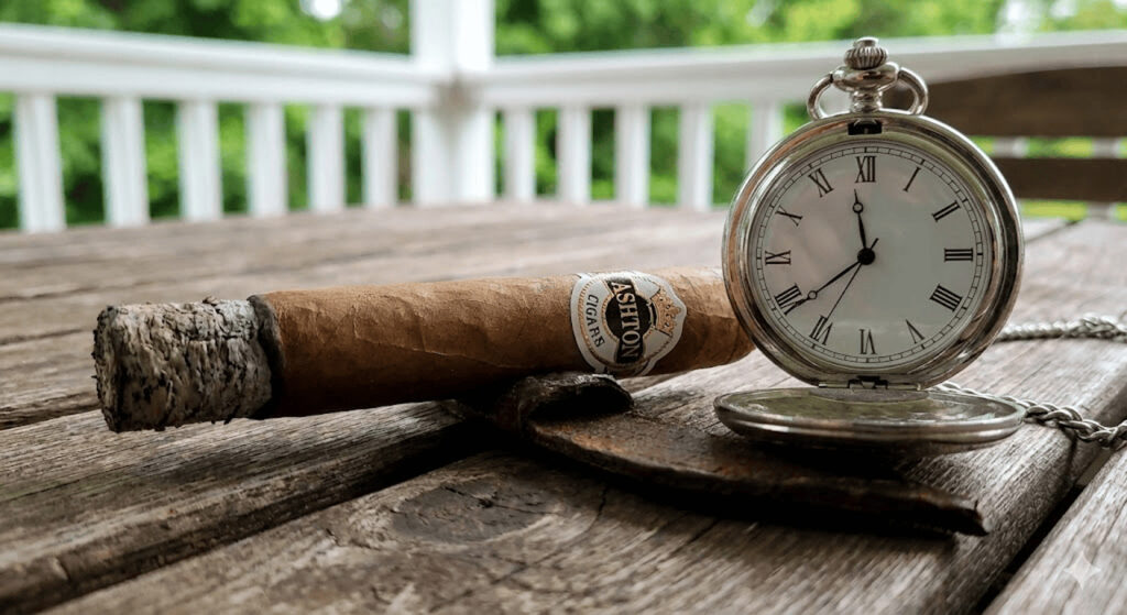 Close-up of premium Ashton cigar resting beside vintage pocket watch on wooden porch table, timeless luxury at our cigar shop Broken Arrow.