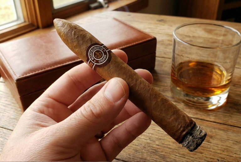 Hand holding a lit Montecristo cigar near a whiskey glass and leather box, perfect for a cigar shop in Broken Arrow.