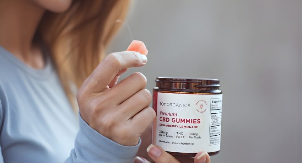 Customer at a CBD shop Tulsa holding a THC-free strawberry lemonade gummy from an organic Joy Organics product jar.