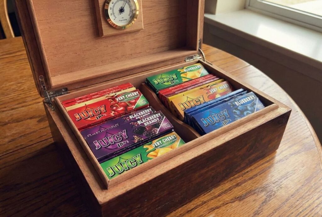 An open wooden humidor filled with an assortment of different flavored Juicy Jay's rolling papers, including Cherry, Blueberry, and Blackberry, illustrating organized storage for rolling papers broken arrow.