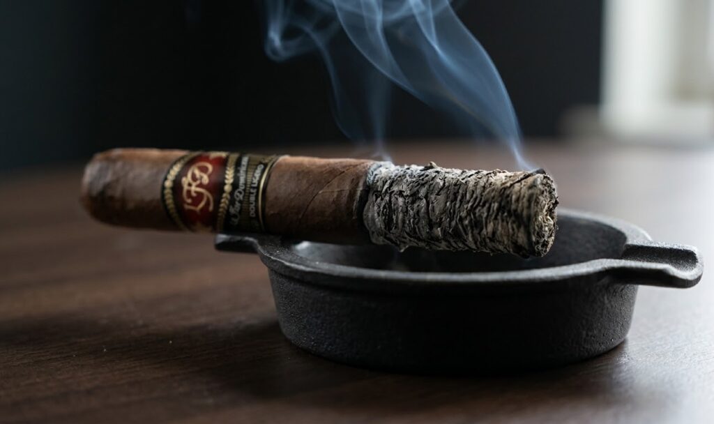 A lit premium cigar rests on a black ashtray, producing blue smoke. Find a wide selection at our local cigar shop tulsa.