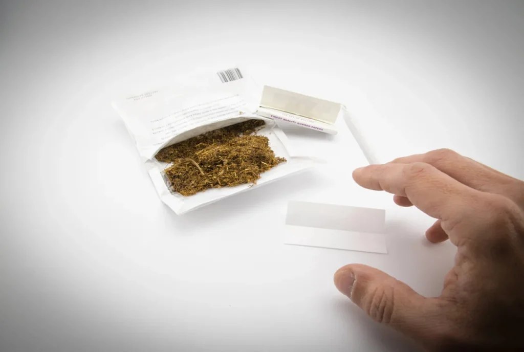 A hand reaching for a fresh sheet of Rolling papers tulsa, positioned next to an open tobacco pouch.