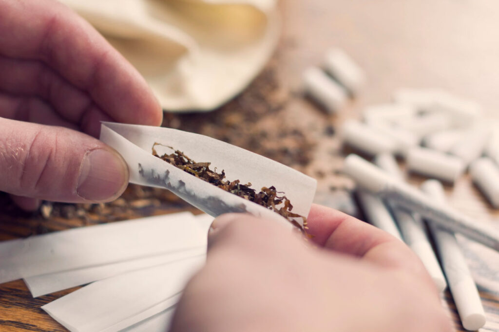 Hands preparing loose leaf tobacco using rolling papers Tulsa style on a wooden table with filters and extra wraps.