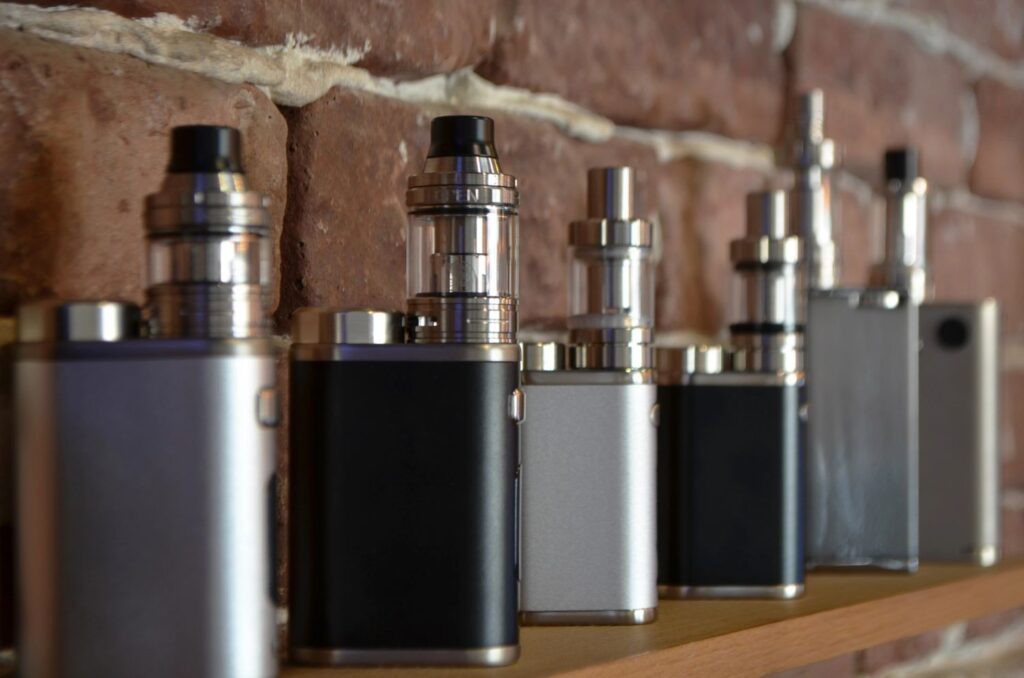 Large collection of the best vape mods Owasso selection on a shelf featuring silver and black box mods with glass tanks.