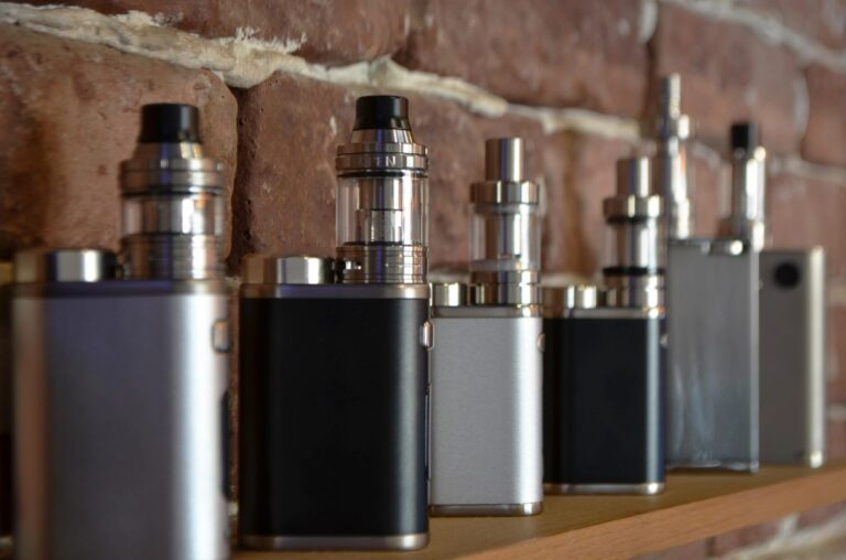 Large collection of the best vape mods Owasso selection on a shelf featuring silver and black box mods with glass tanks.