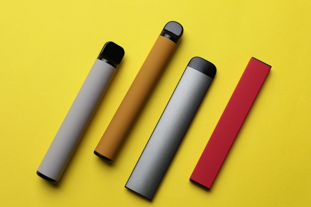 Flat lay of grey, tan, silver, and pink nicotine pods tulsa vaping devices diagonally on a solid yellow background. 