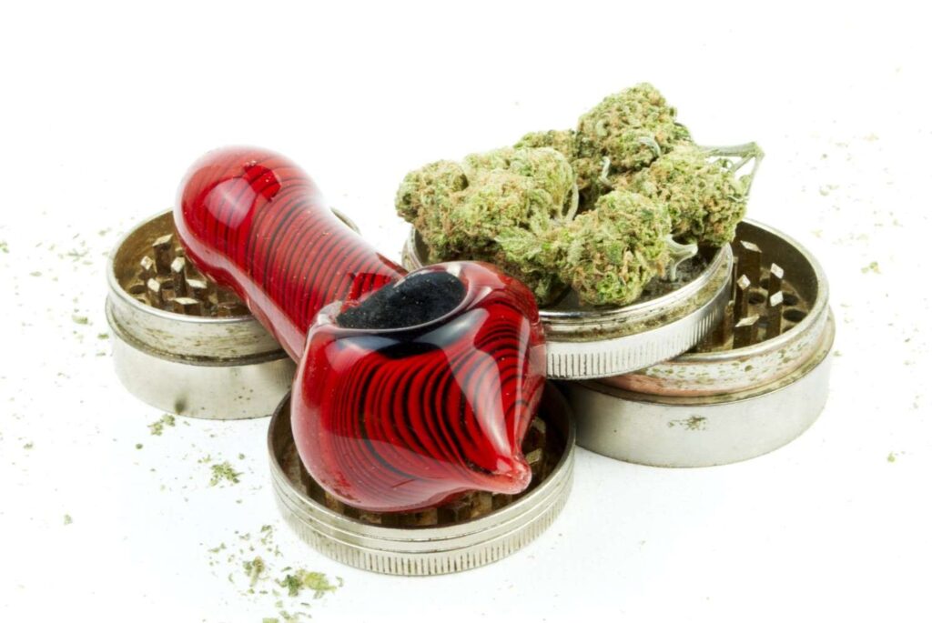 Vibrant red spoon pipe from a glass pipes Tulsa retailer resting on a disassembled metal herb grinder with buds. 