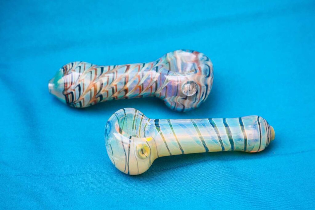 Two colorful custom spoon style glass pipes Tulsa glassblowing art displays with intricate swirl patterns on blue fabric 