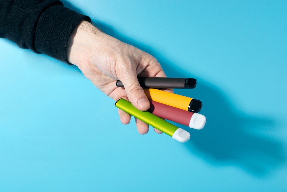 A hand holds four colorful disposable vape pens over a blue surface, showcasing products from a top smoke store broken arrow.