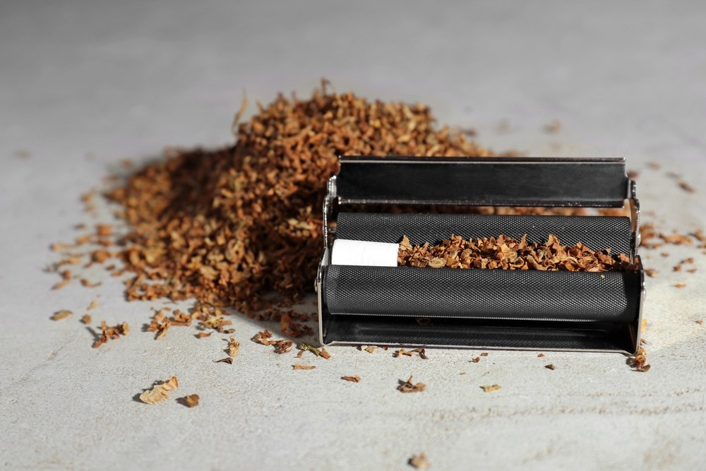 Loose tobacco pile next to a metal cigarette roller on a grey concrete surface. High-quality rolling papers sapulpa review.