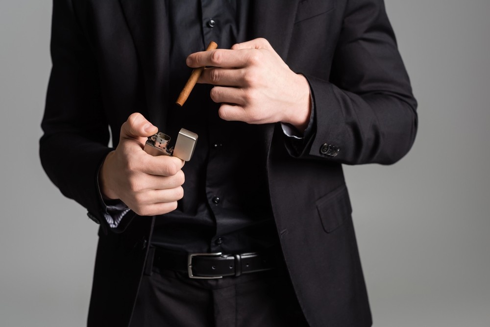 Man in a black suit and shirt holds an open silver lighter and a premium cigar, ready to smoke near our cigar store Owasso.