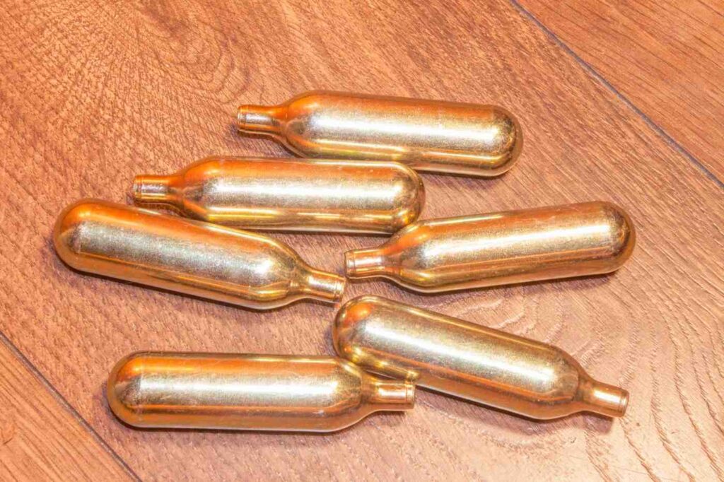 Six gold nitrous oxide Owasso whip cream chargers scattered in a loose pile across a polished brown wood floor.
