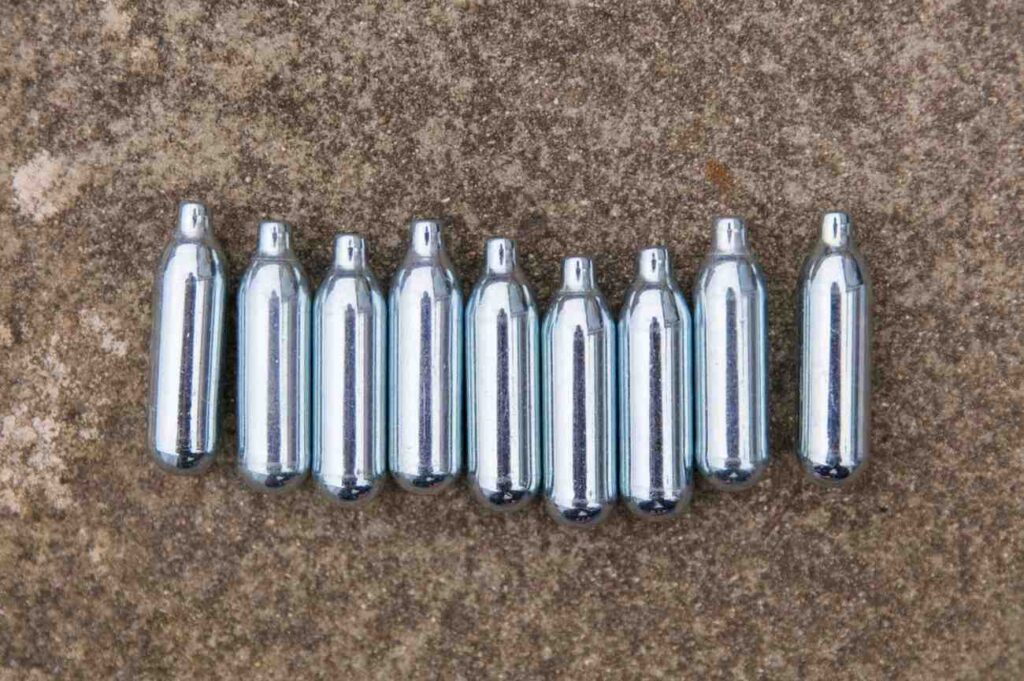 Nine metallic silver nitrous oxide Owasso culinary gas canisters arranged in a neat horizontal row on concrete. 