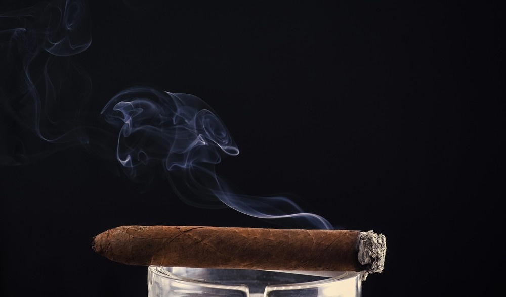 A premium lit cigar rests on a crystal ashtray with elegant wisps of smoke, showcasing stock from our cigar store Owasso.