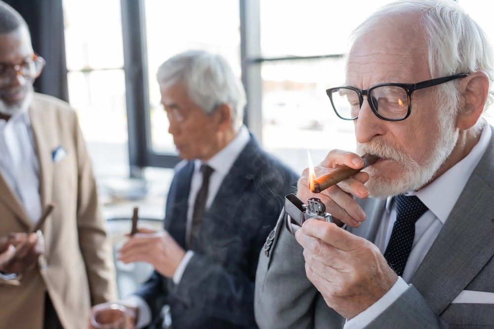 A senior man in a suit lights a premium cigar using a metal lighter at a gathering, representing our cigar store Owasso.