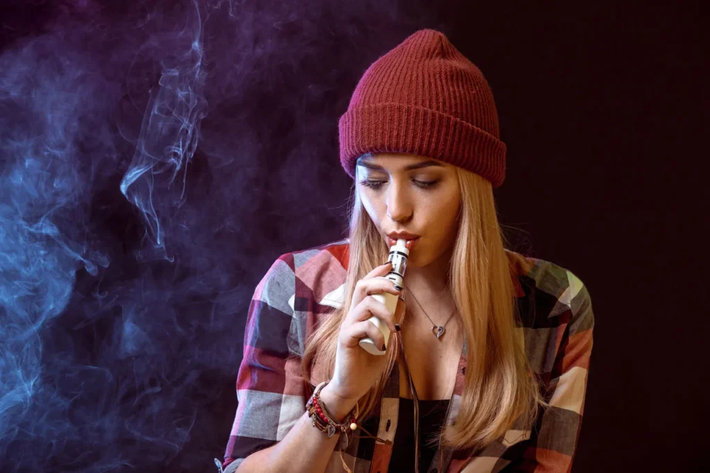 A woman in a red beanie using a white mod vaporizer, showcasing smoke accessories Tulsa for local vape shop patrons.