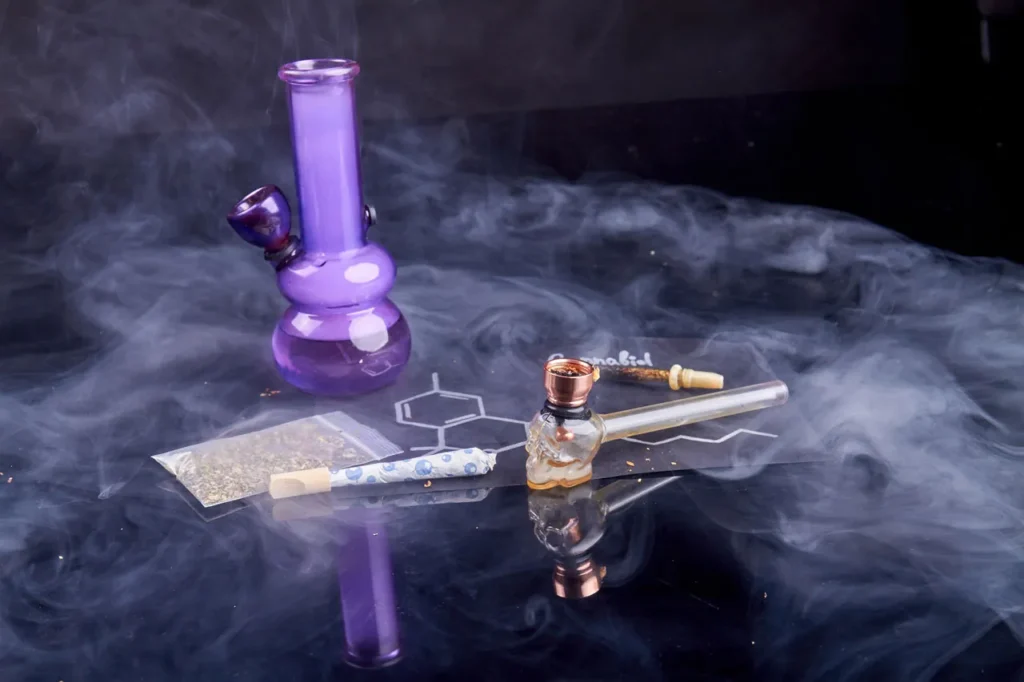 Variety of smoke accessories Tulsa including purple glass bong, skull pipe, and pre-roll on a smoky black background.