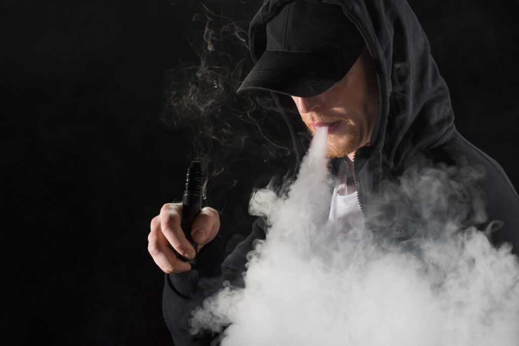 Man in black hoodie using powerful vape mods Owasso for cloud chasing against a dark background with thick vapor clouds. 