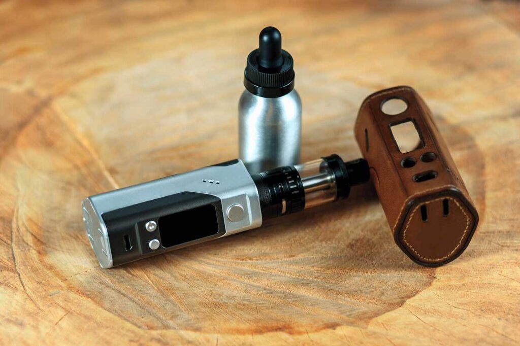 A silver box vape mod with a black tank next to a leather protective case and an e-liquid bottle from vape mods Tulsa.