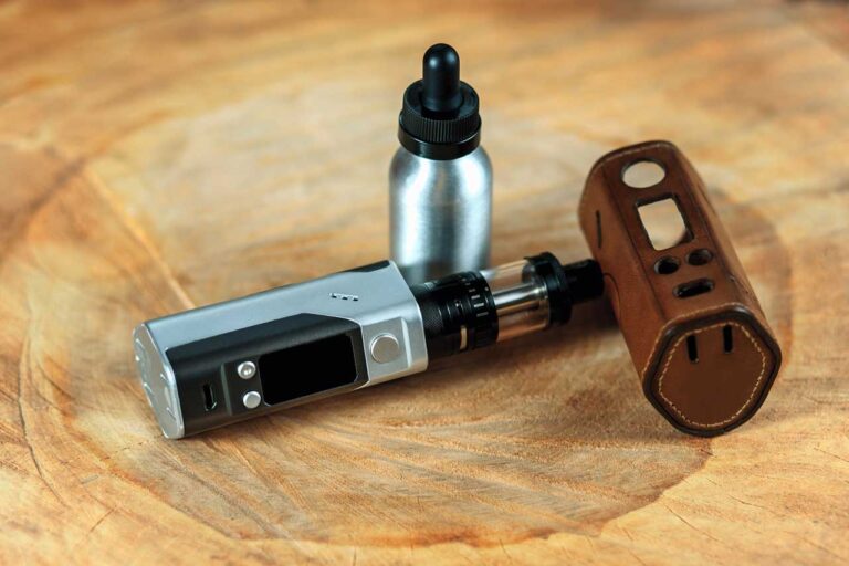 A silver box vape mod with a black tank next to a leather protective case and an e-liquid bottle from vape mods Tulsa.